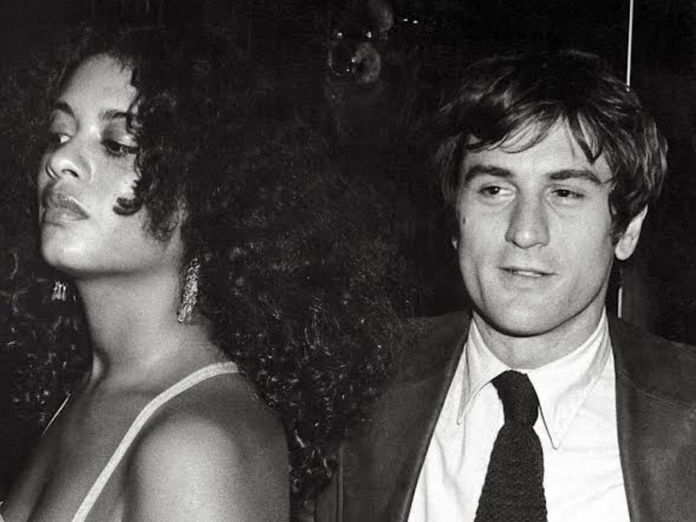 Robert De Niro e sua primeira esposa, a atriz Diahnne Abbott. A foto em preto e branco mostra uma mulher negra, de cabelos longos e cacheados ao lado de um homem branco, usando terno 