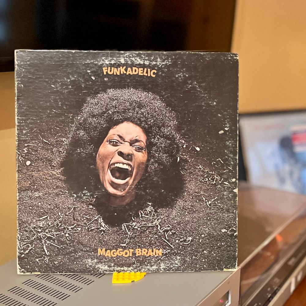 The cover of the 1971 album by the band Funkadelic, titled Maggot Brain. The album art features the screaming face of a dark-skinned woman, submerged from the neck down in dark, barren soil, representing a distressed “Mother Earth”.