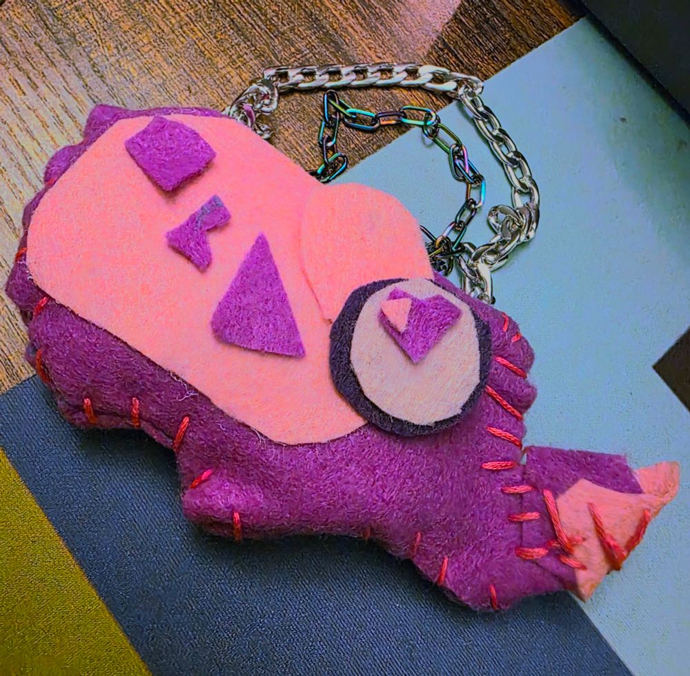 A beetle made out of four different colours of felt ranging from a purple body to pink wings and horn. It is sewn together with hot pink embroidery thread, and has silver and anodized metal link chains attached to the top of it, for hanging up.

It has a heart shaped pupil and is named Lovesick!