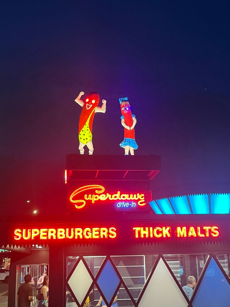 The outside of the Superdawg drive-in. On top of the building are two anthropomorphized hot dogs. One of them has eyes that glow in a menacing shade of red, the other has eyes that glow in an eerie shade of blue.