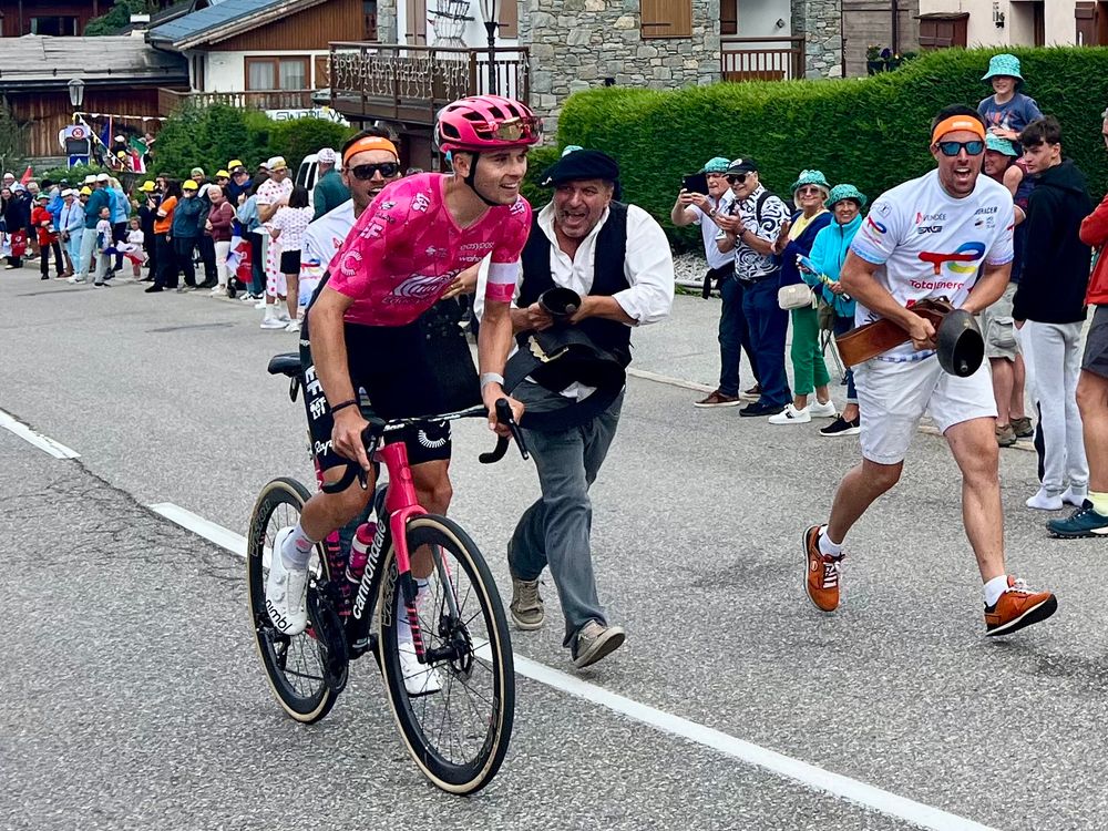 Man in beret ringing a cow bell while a bike rider makes his way uphill 