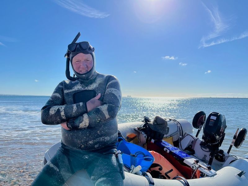 Smiling Eric Smith in diving gear leaning on a boat on the beach on a sunny morning