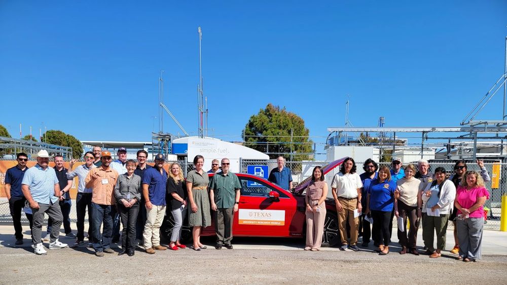 Outdoor group photo taken in front of a red hydrogen-powered vehicle for a workshop highlighting unique characteristics of hydrogen. The photo captures a deep blue cloudless sky and 15-20 smiling people. 