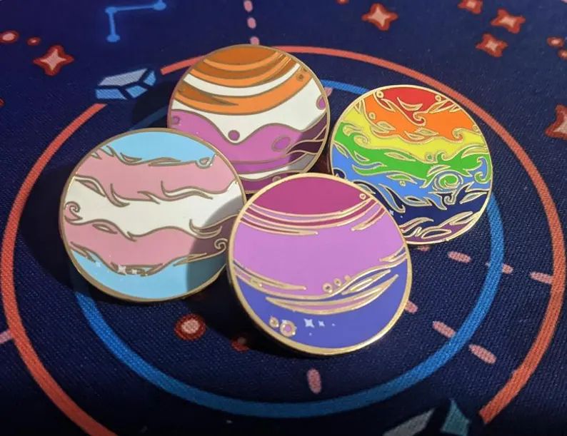 lgbt planet enamel pins! center bottom is the BiSexual flag, left is the Trans flag, top is the Lesbian Flag and to the right is the Rainbow (or Gay flag). Hard enamel, gold pins designed by Kaiya Kaiju Art!