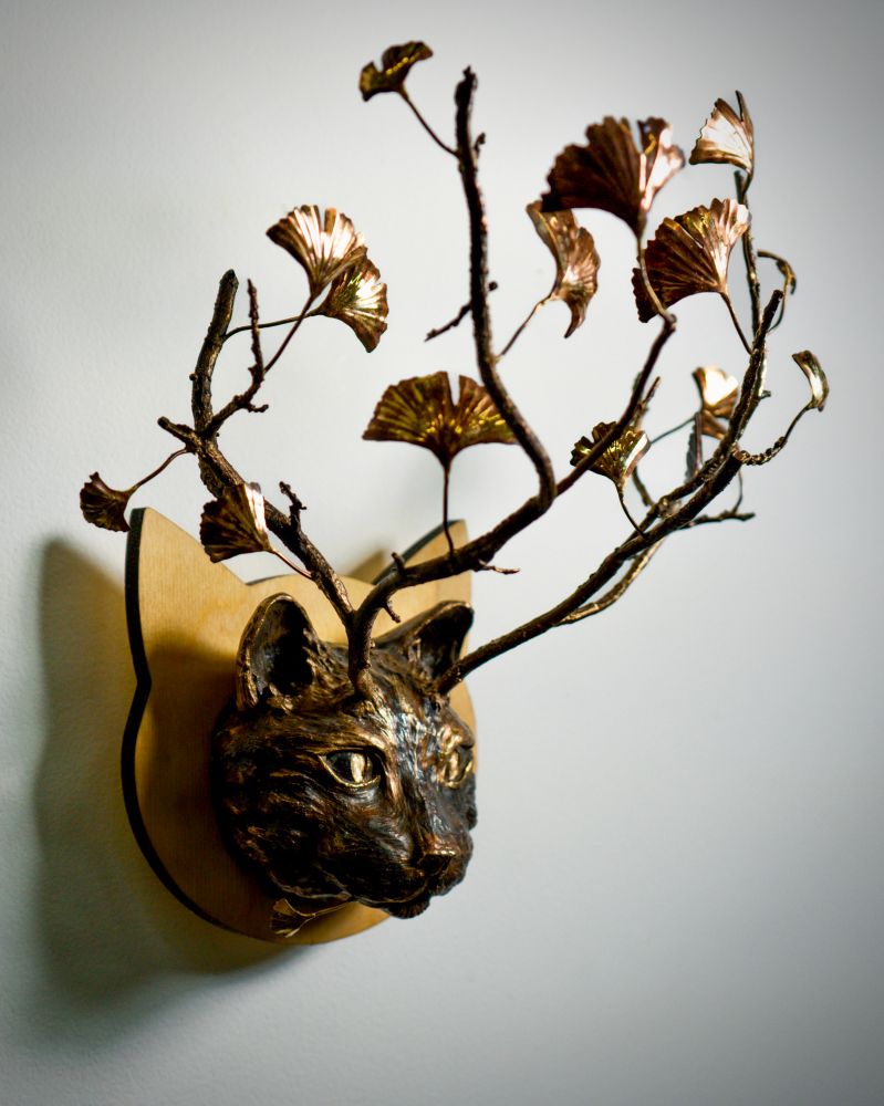 Three-quarter profile of a bronze wildcat head with branch antlers and ginkgo leaves. The cat head is mounted on a light wooden panel. 