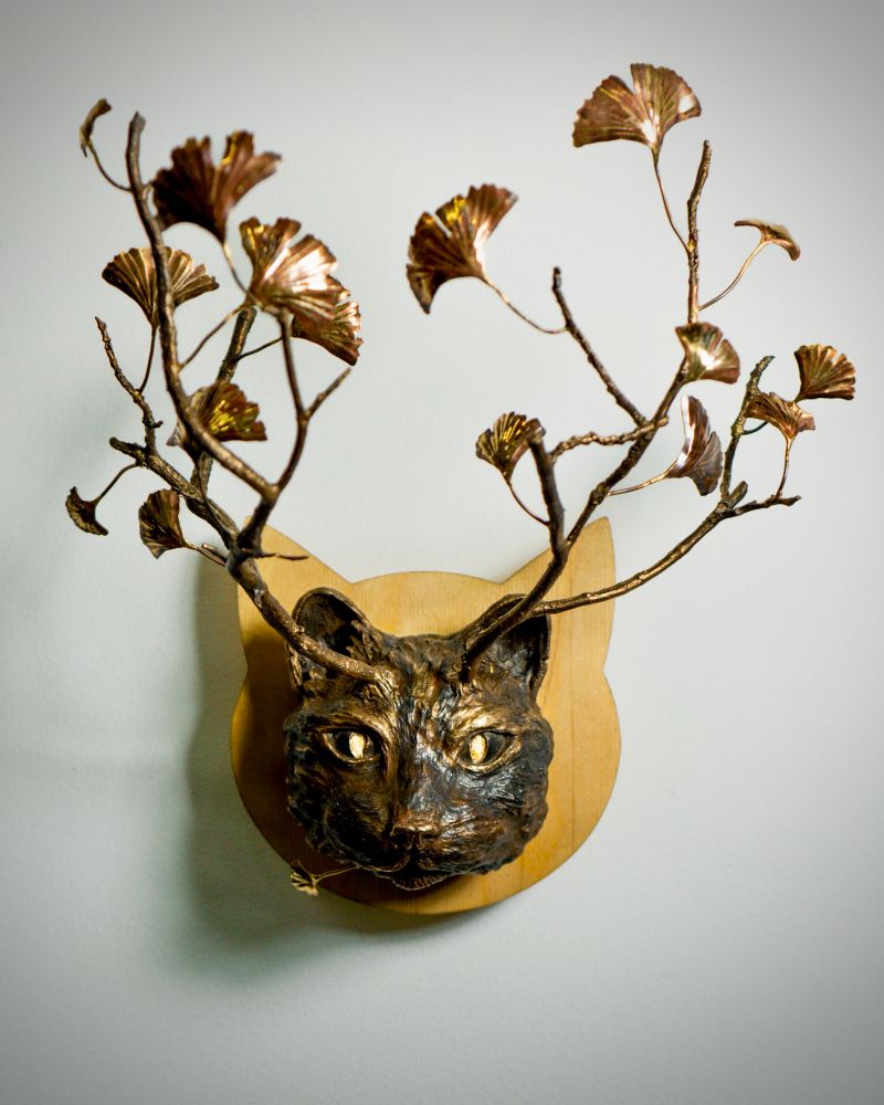 Direct view of a bronze wildcat head with branch antlers and ginkgo leaves. The cat head is mounted on a light wooden panel. 