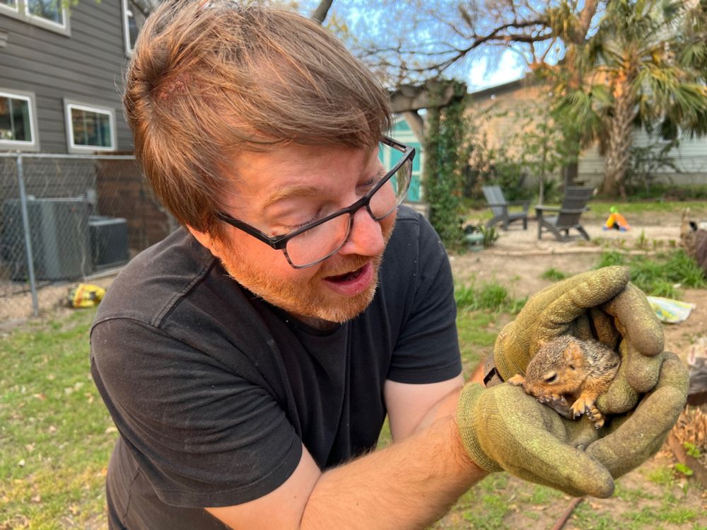 Bespectacled white man in awe and holding a young squirrel in a backyard 