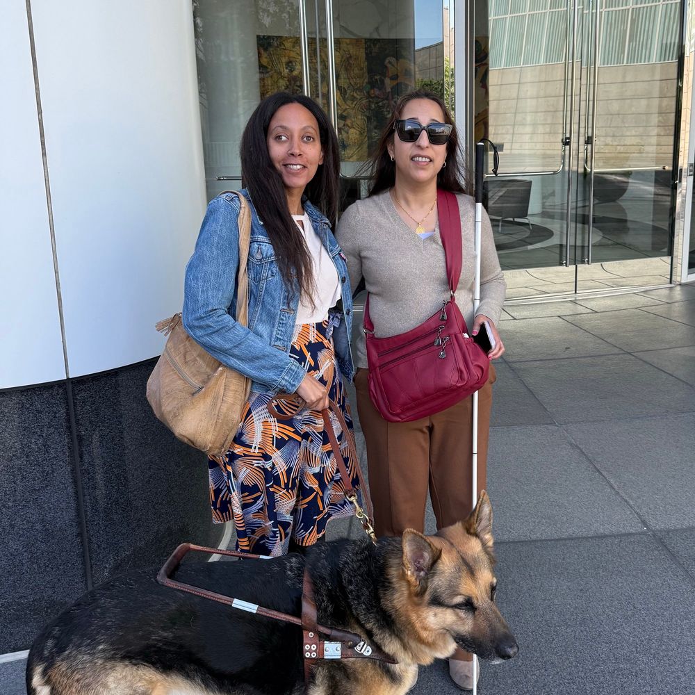 Two women of color stand together smiling. Deepa wears a gold necklace over a gray top, dark yellow trousers, sunglasses, and holds her cane and iPhone in her left hand. Haben wears a denim jacket over a colorful dress and holds the leash of her Seeing Eye dog in her right hand. Behind them is a glass door reflecting the trees in front of the building.