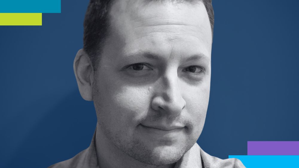 Alt text: A close-up portrait of a man with short, dark brown hair, slight stubble, and light skin. He is wearing a light blue collared shirt and has a confident, subtle smile. His eyes are looking directly at the camera. The background is a plain, off-white wall with soft lighting creating a gentle shadow on the left side of his face.