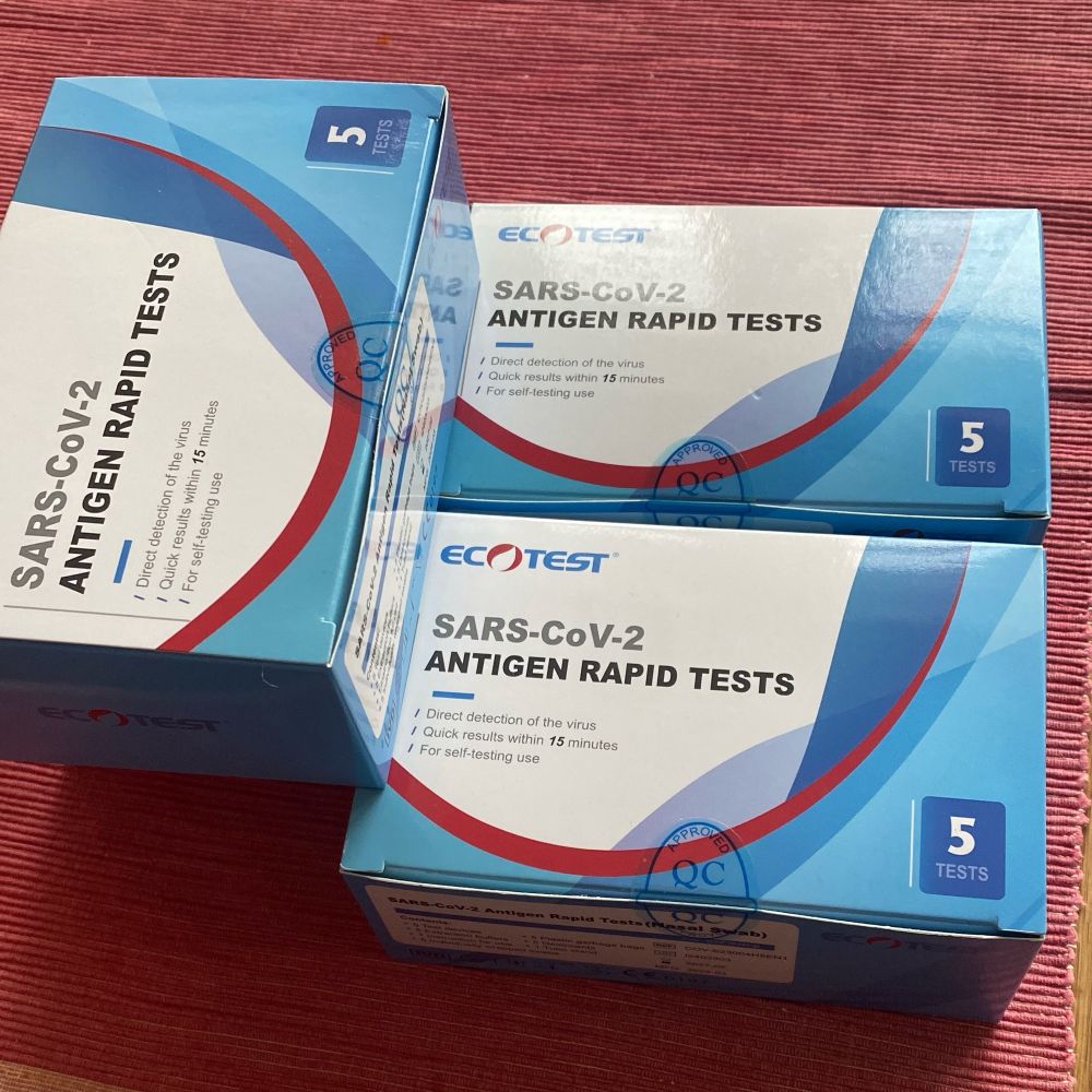 4 boxes of 5 each Covid RAT tests. The boxes are mostly blue, and they’re on a brick-red corrugated cotton placemat
