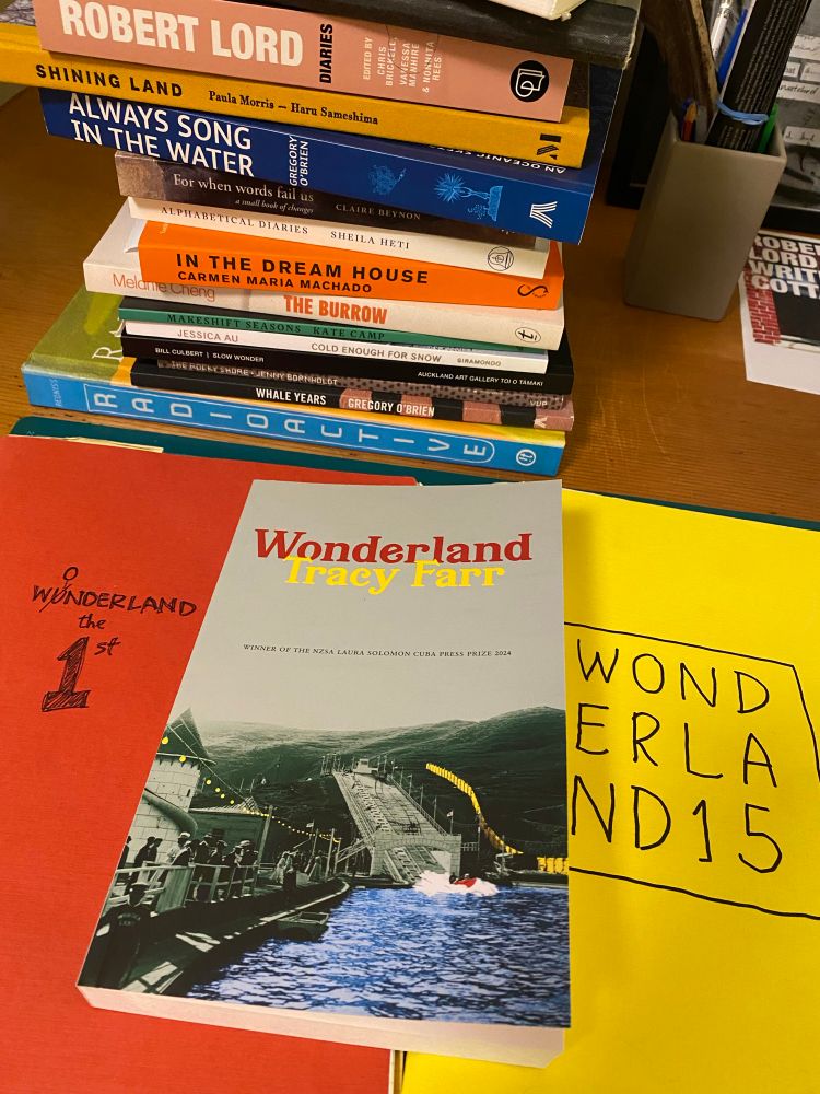 A novel with WONDERLAND in red text and Tracy Farr in yellow text. There are two notebooks underneath it, one red (Wonderland the 1st), the other yellow (Wonderland 15).
