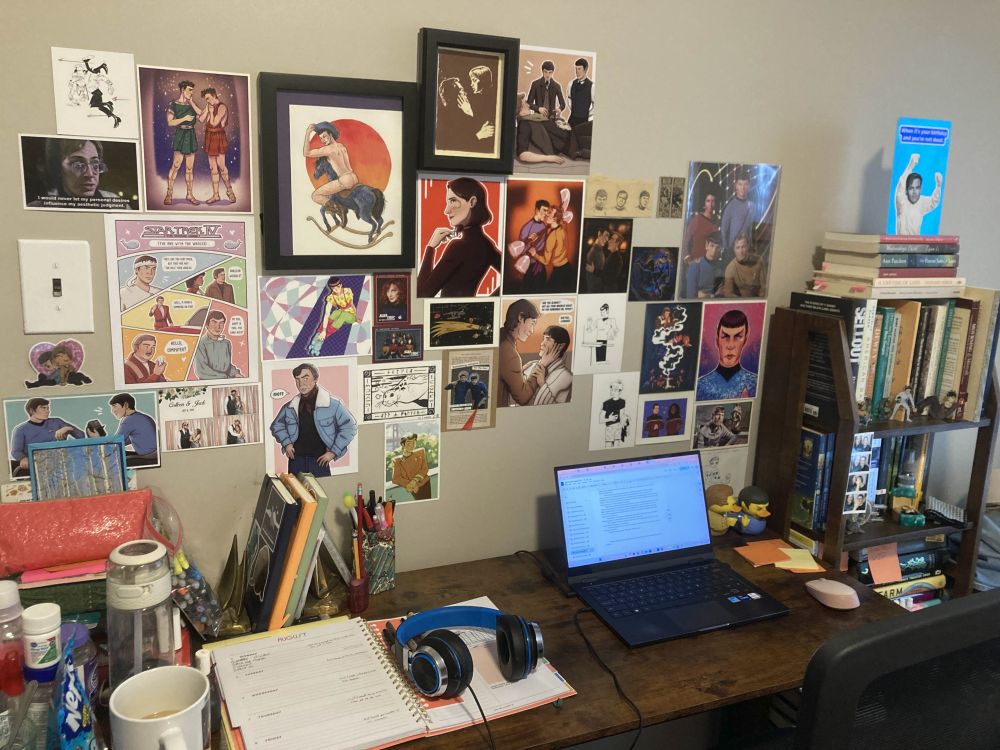 A writing desk. A laptop is open to a Google doc and to the left is an open agenda with headphones resting on top of it. On the wall is a collection of different Star Trek art, some prints and some original paintings. To the right of the desk is a bookshelf