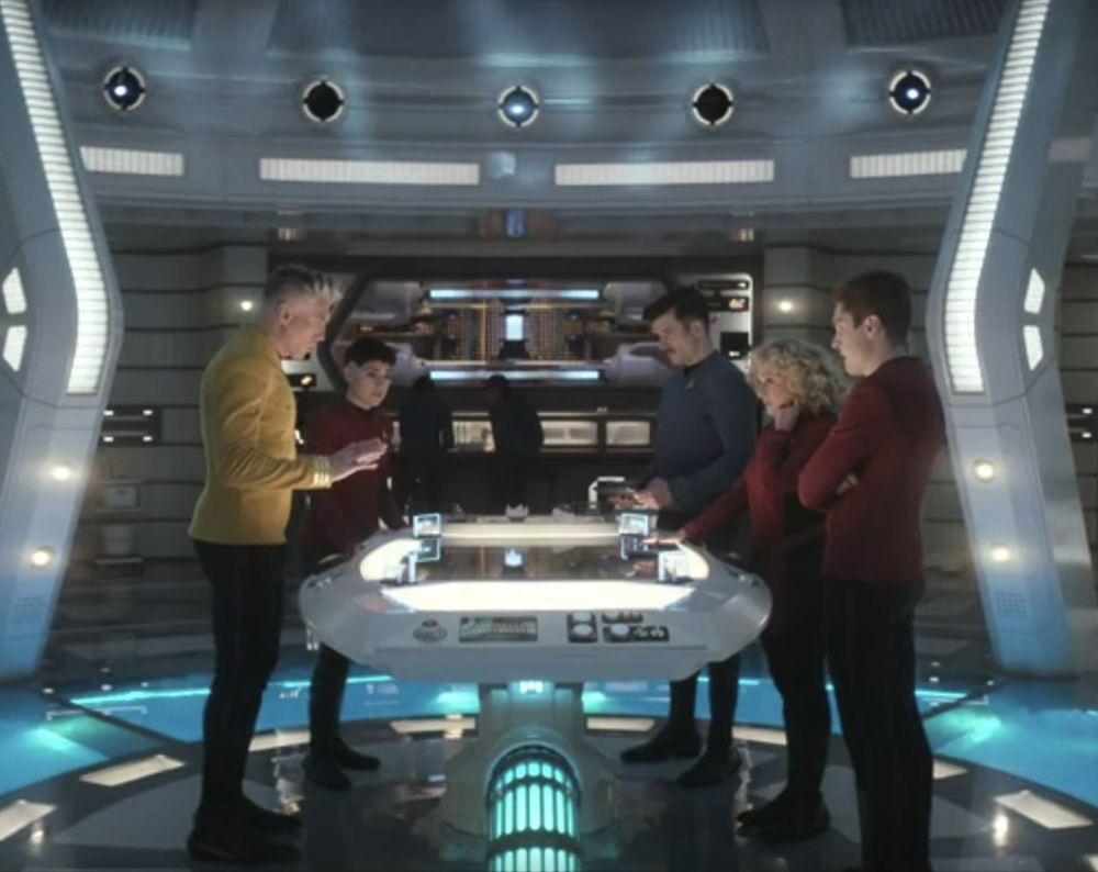 A screenshot from Season 3 Episode 5 "Through the Lense of Time" of Star Trek: Strange New Worlds. Captain Pike, Ortegas, Sam Kirk, Pelia, and Scotty are all standing around a table having a deep discussion.