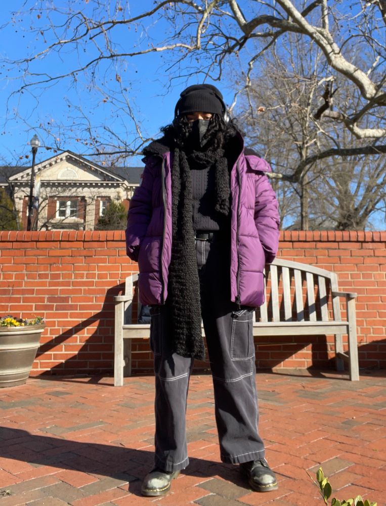 A photo of the same outfit conceptualized now worn in real life, copying the same pose as the doodle. OP wears a black beanie, headphones, a N95 black mask, a long unzipped plum colored puffer jacket, wide leg black pants, and boots. The unzipped jacket reveals the rest of the outfit: a long black knitted scarf, a cable knit sweater tucked into the pants with a black leather belt, and black leather gloves. Op stares at the camera, his eyes the only thing visible between his hat, his mask, and his locs. 