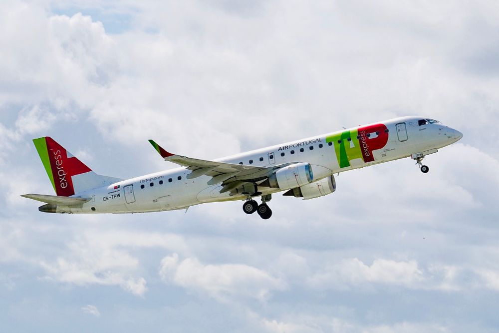 TAP Express Embraer E190 aircraft, registration CS-TPW, taking off under partly cloudy skies. The plane features the vibrant red, green, and white TAP Air Portugal livery, with landing gear retracting as it climbs into the sky.