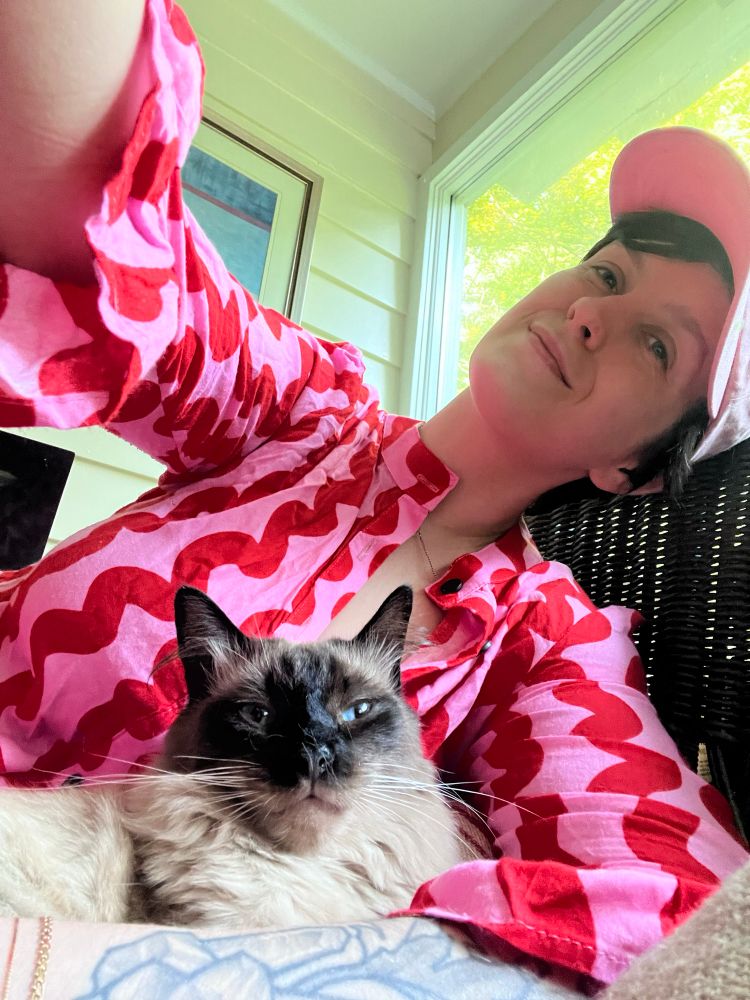 Selfie of Aliya with her cat, Jeremy. Aliya is wearing a pink and red dress and a pink cap. She and Jeremy are sitting on a wicker couch and her arm is around him. He is a Balinese cat (color point with light body and a dark face, ears, tail, and paws). They both look calm.