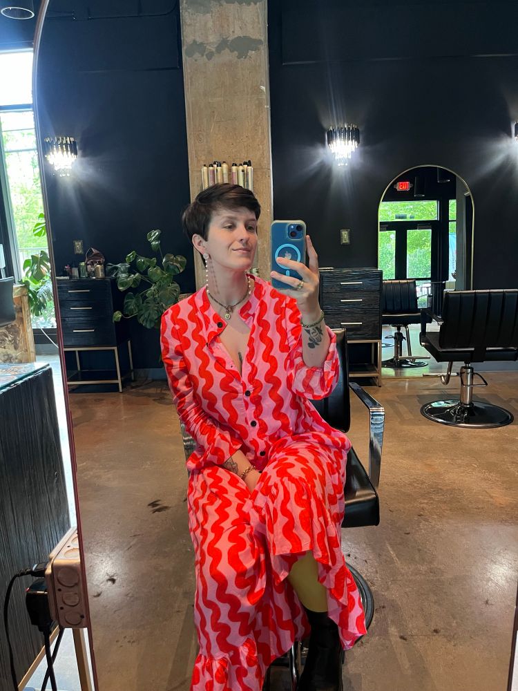 Mirror selfie of Aliya post-haircut. She is in a salon, seated at the mirror, wearing a pink maxi dress with vertical red squiggles (she loves pink and red together). She looks so good y’all. Like wow.