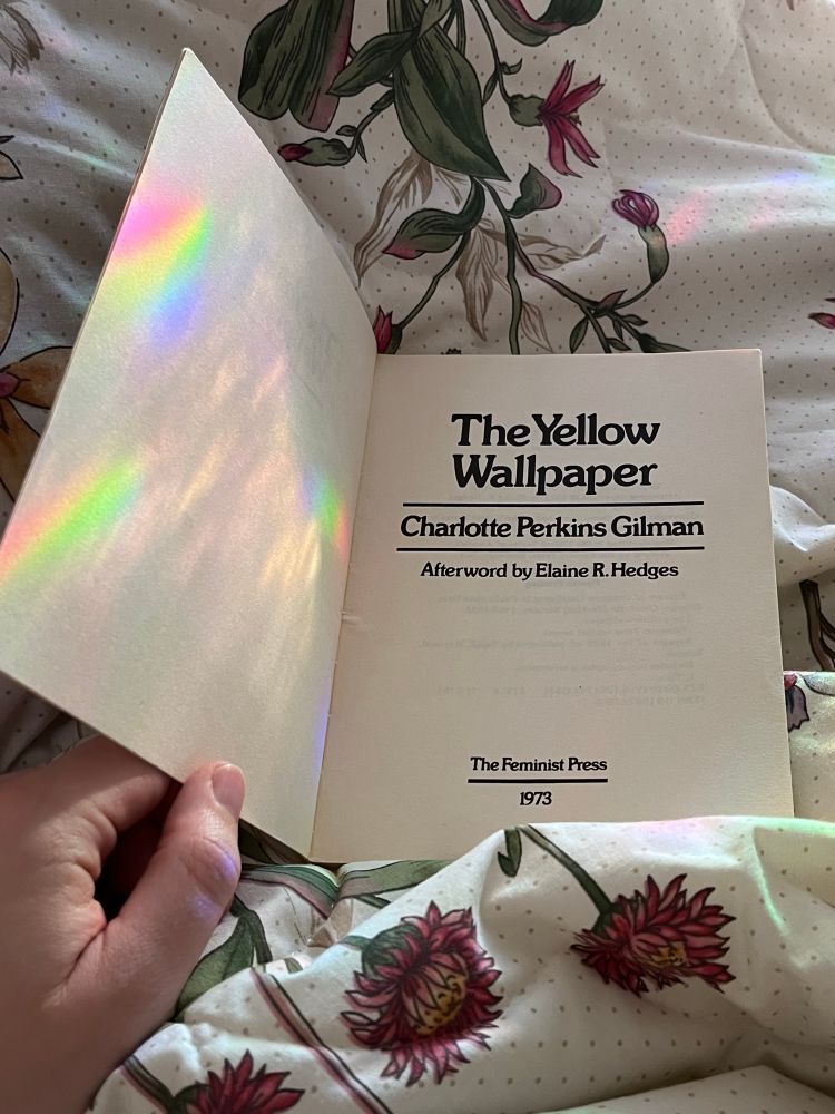 A photo of a hand holding the book The Yellow Wallpaper open to the title page. It is lying on a blanket with a floral print and there is a rainbow cast onto lefthand side of the book from nearby window light (due to a privacy film meant to cast rainbows).