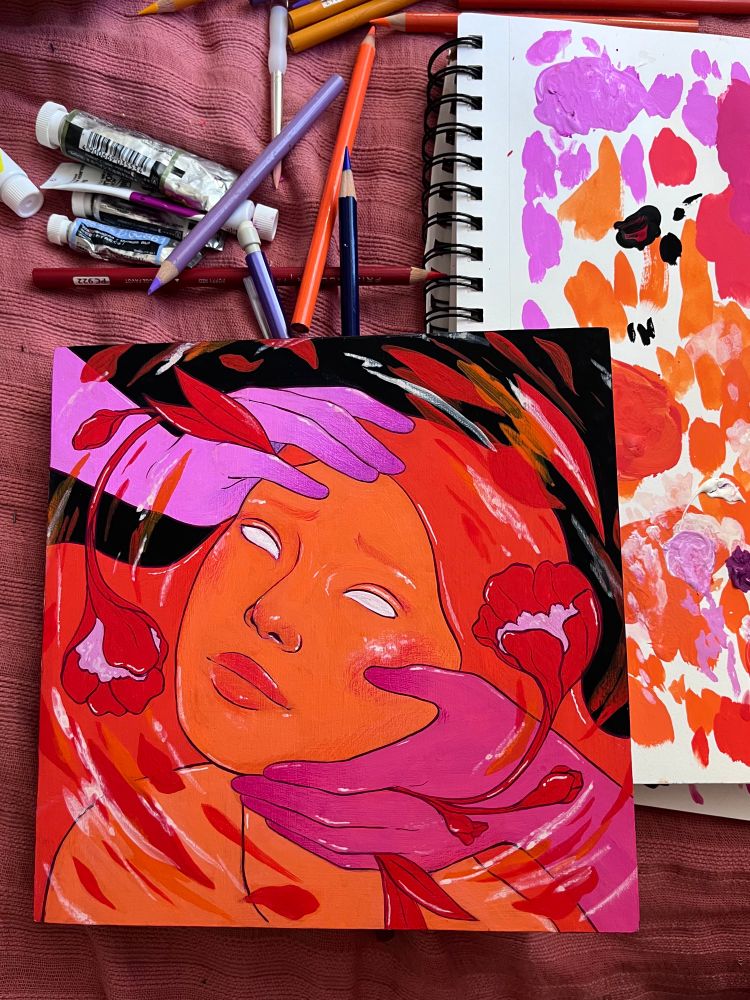Bird’s eye view of a square painting, an open sketchbook, and a slew of tubes of gouache and colored pencils all on a pink blanket. The painting is of two hands (one light-medium purple, one sort of dark pink) coming from opposing corners and holding the face of a woman looking up. The woman is painted orange with bright red-orange hair. She does not have pupils or irises (this is very common of my work). There are red flowers growing out from where the hands are touching the face. And there is a ripple effect around the face of brush strokes and implied petals and leaves. There is a black background behind all of this. The open sketchbook shows it has been used as a palette for the gouache.