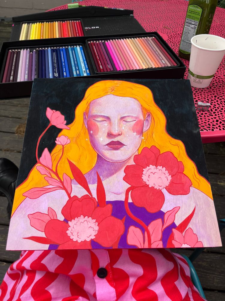 Photo of one of Aliya’s mixed media pieces on a wood panel. The piece is sitting partly on her lap and partly leaned against a pink metal outdoor table. It features a feminine figure with light purple skin and vibrant yellow-orange hair and there are red/pink flowers growing out of her chest. On the table behind, there is a set of Prismacolor colored pencils, a white paper cup, and a bottle of green juice. 