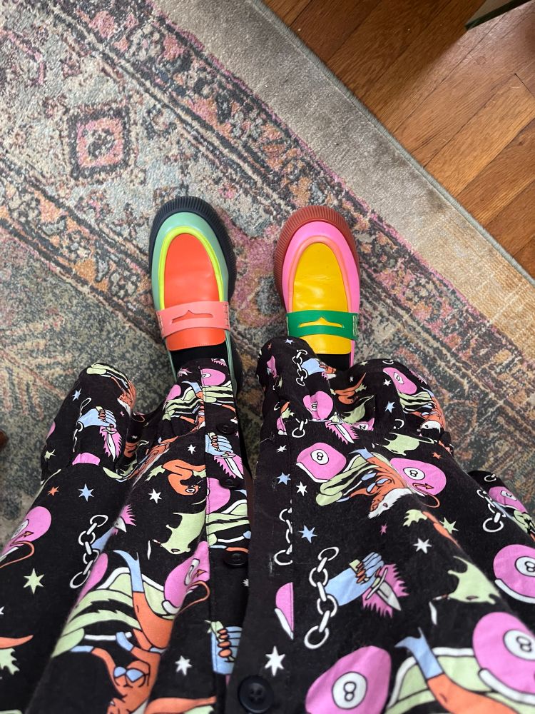 A photo looking down at Aliya’s shoes and the bottom third of her maxi dress. The shoes are (intentionally) mismatched loafers. The left shoe is orange, coral, highlighter yellow/green, mint, and has a black sole. The right shoe is yellow, medium green, coral, bubblegum pink, and has a brown sole. The dress is black with a print featuring a woman in a bikini riding a moped but the wheels are pink magic 8 balls. There are also hands holding knives and rats smoking cigarettes in the print lol.