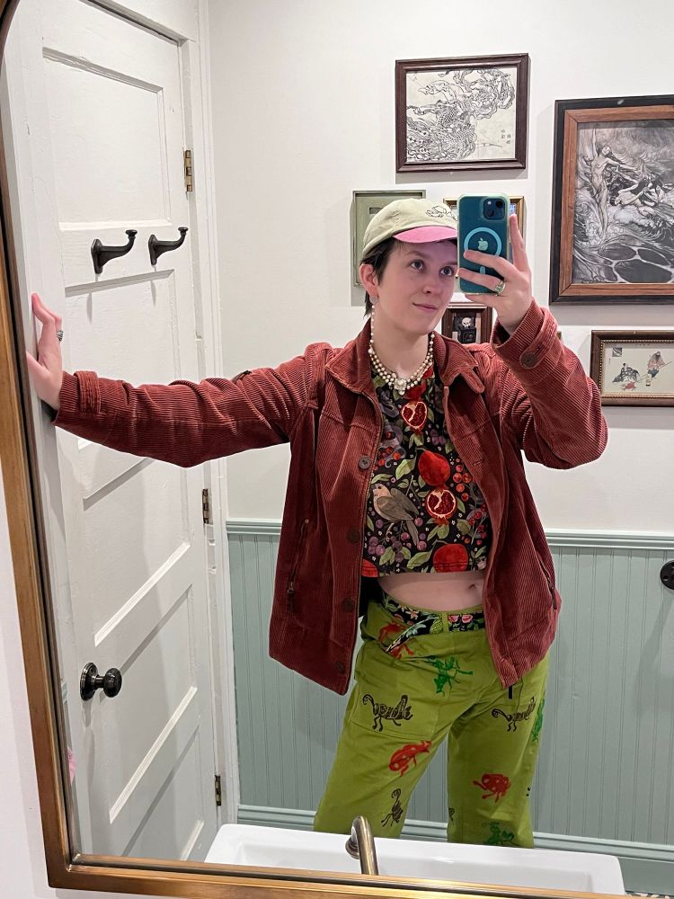 Mirror selfie (Aliya) showing off an outfit.
She is wearing:
- A light, muted green and muted rosy color cap
- A crop top from Morning Witch with a black background and large print of pomegranates, snakes, and doves
- Chartreuse pants from Big Bud Press with a hand stamped print of ladybugs, grasshoppers, and praying mantises. The print is very spread out
- A floral belt with a black background
- A muted deep rosy corduroy jacket
- Lots of jewelry, including a pearl necklace and dangly pearl earrings

She is in a bathroom with a gallery wall behind her.