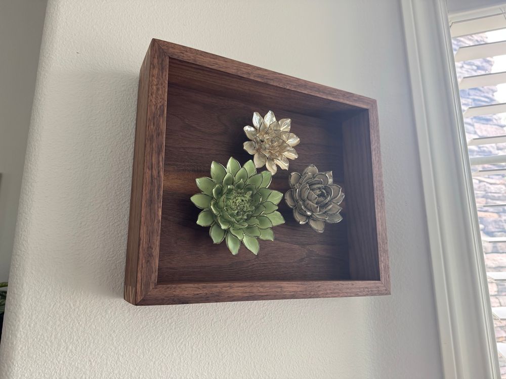 Walnut shadow box with three ceramic flowers in the center. Maple splines in the mitered corners forming a crisscross pattern.