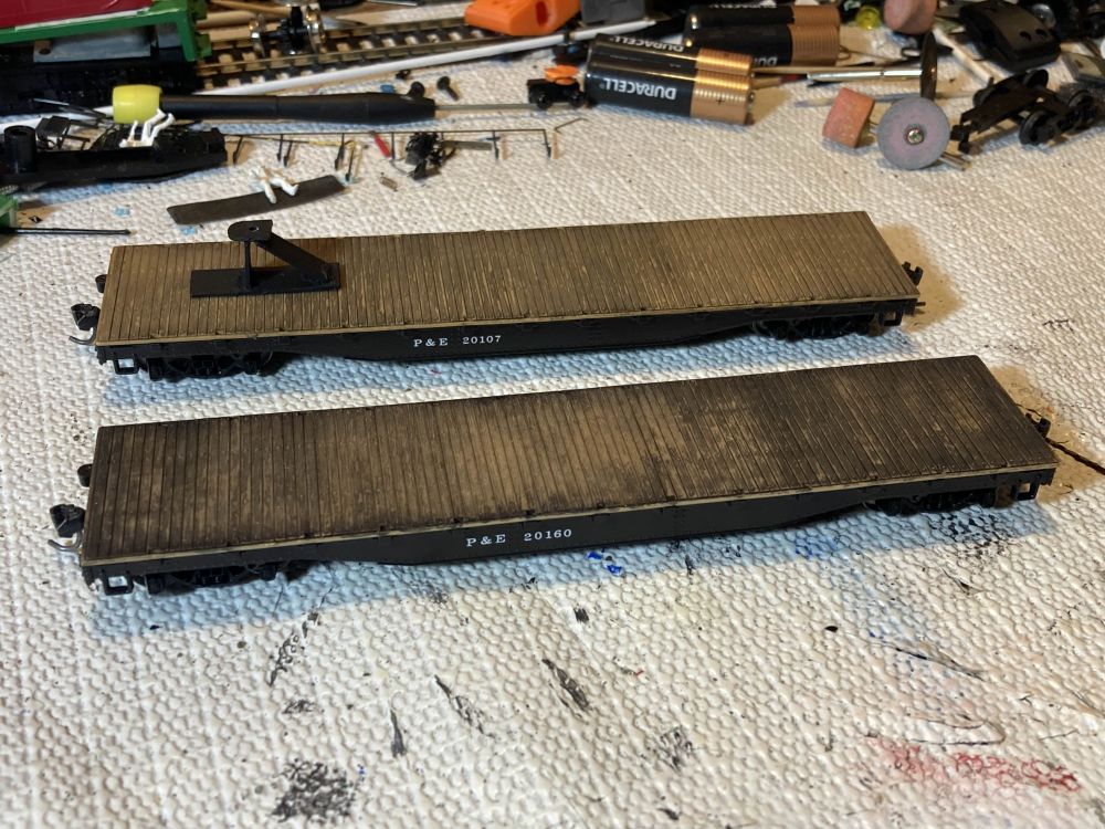 Two models of flatcars sit on a workbench. The further one (P&E #20107) has less weathering on its deck (and notably a trailer hitch) while the near one (P&E #20160) has more. The P&E is my freelance railroad.
