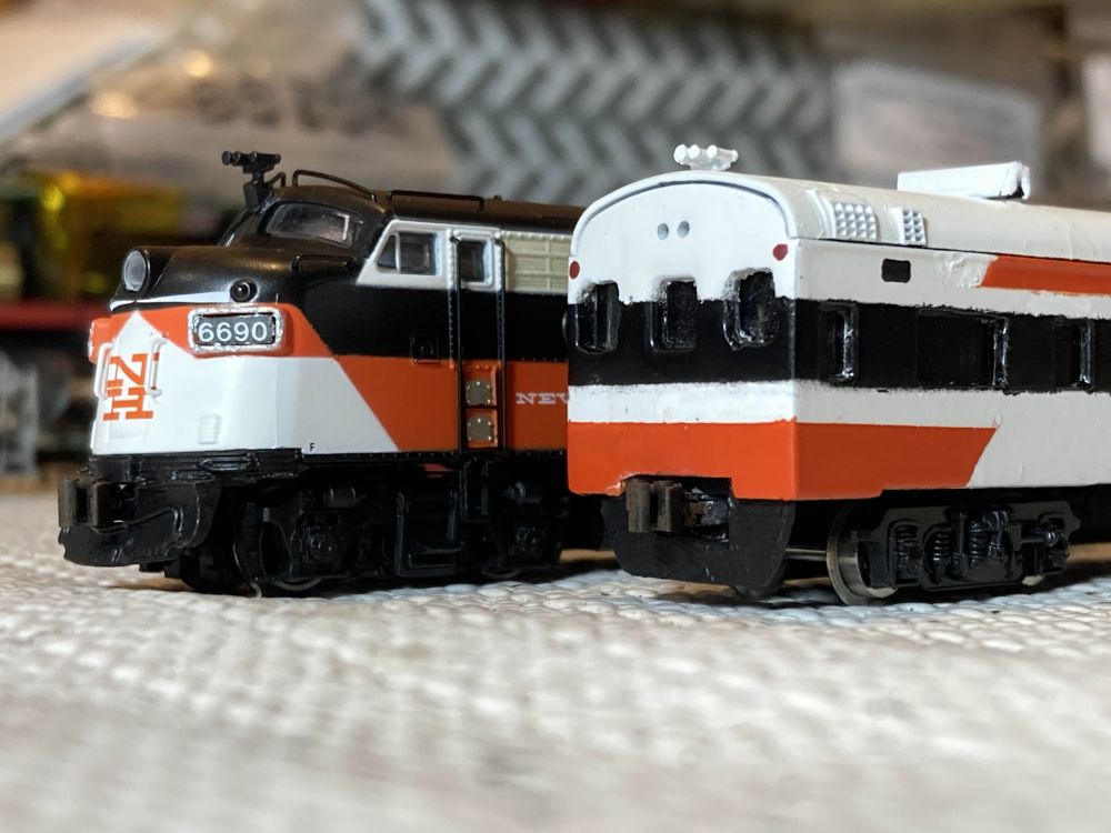 A closeup of two N scale model trains - one a locomotive and the other a cab car. The cab car is an extensively modified coach; the diaphragm has been removed and cab windows are carved into the end. The locomotive has also been modified, with a new plow and horn.