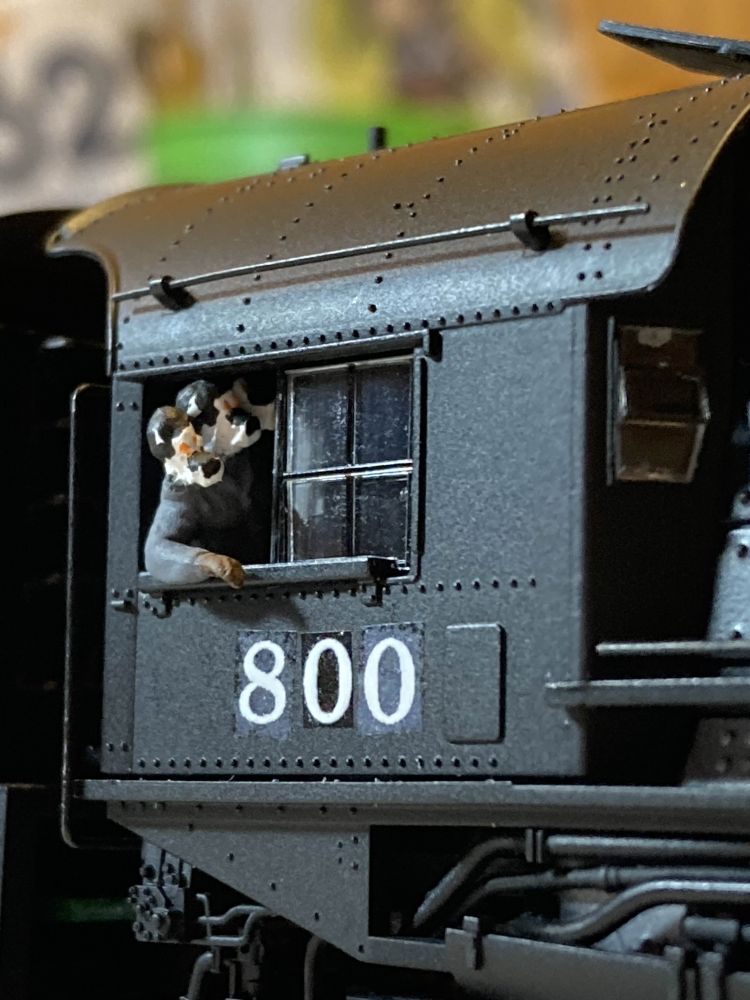 A closeup of a locomotive cab. A figure sticks out of the window with an arm resting on the sill. The figure has been modified to represent myself, Duke the cerberus, with heads from two more figures glued onto the shoulders of the base and snouts added to all three. The heads are mostly white with black "hair" and tops of snouts, with orange eyes painted onto each head. The figure is wearing a gray coat and brown gloves.