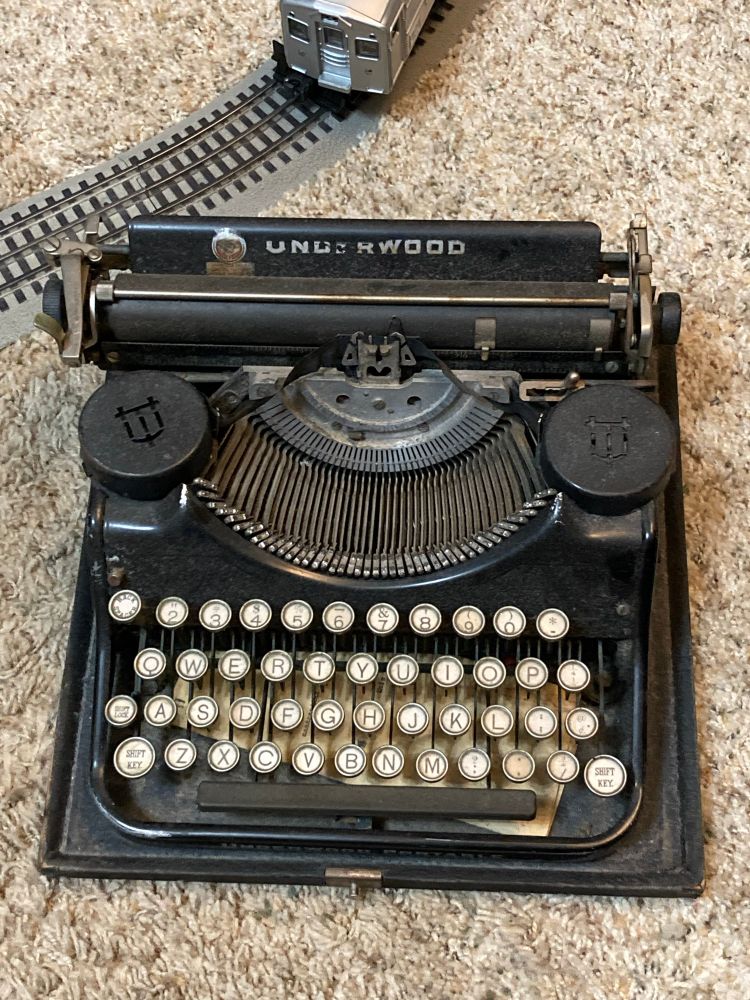 An Underwood portable typewriter, viewed from above, sits on a carpeted floor. It’s rather dusty from storage and neglect but is mostly whole.