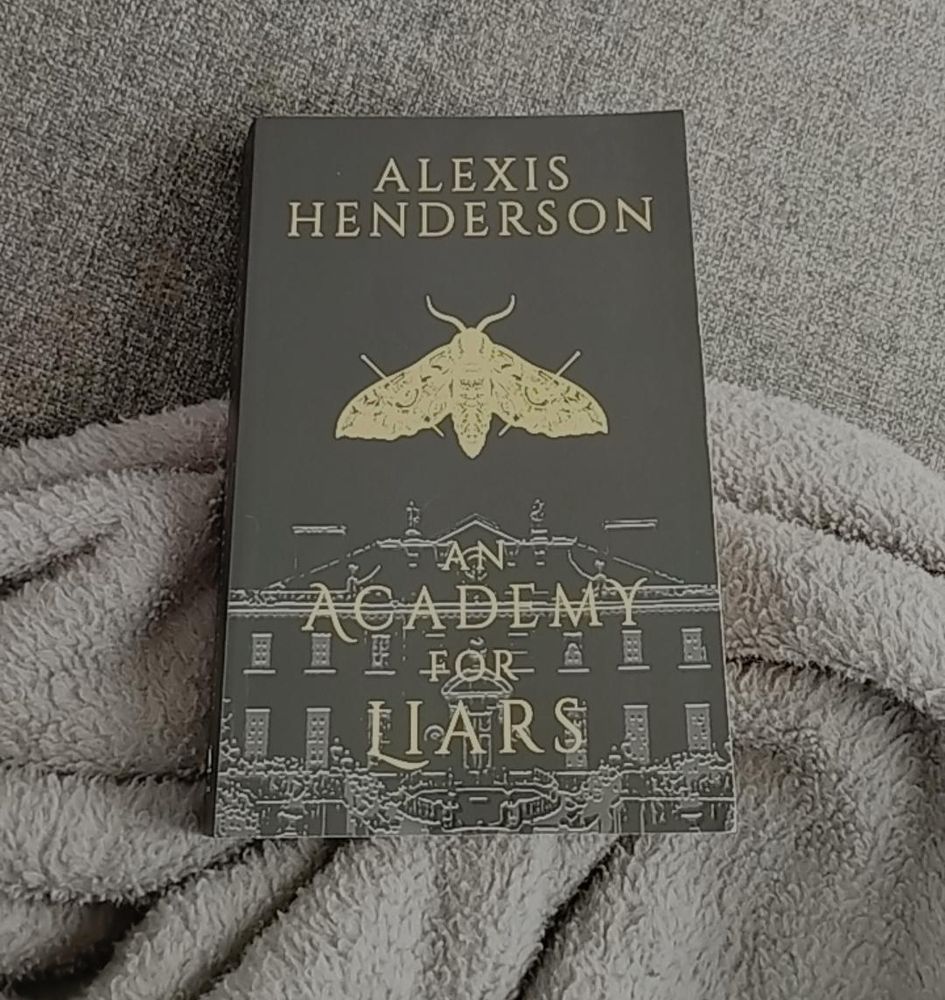 A photograph of the book An Academy by Alexis Henderson laying half on a grey fluffy blanket, half on a grey sofa. The cover of the book is black with a gray illustration of a university style building at the bottom with the title overlayed in gold text. In the middle of the cover is a gold moth with its wings pinioned under the top is the author's name in the same gold. 