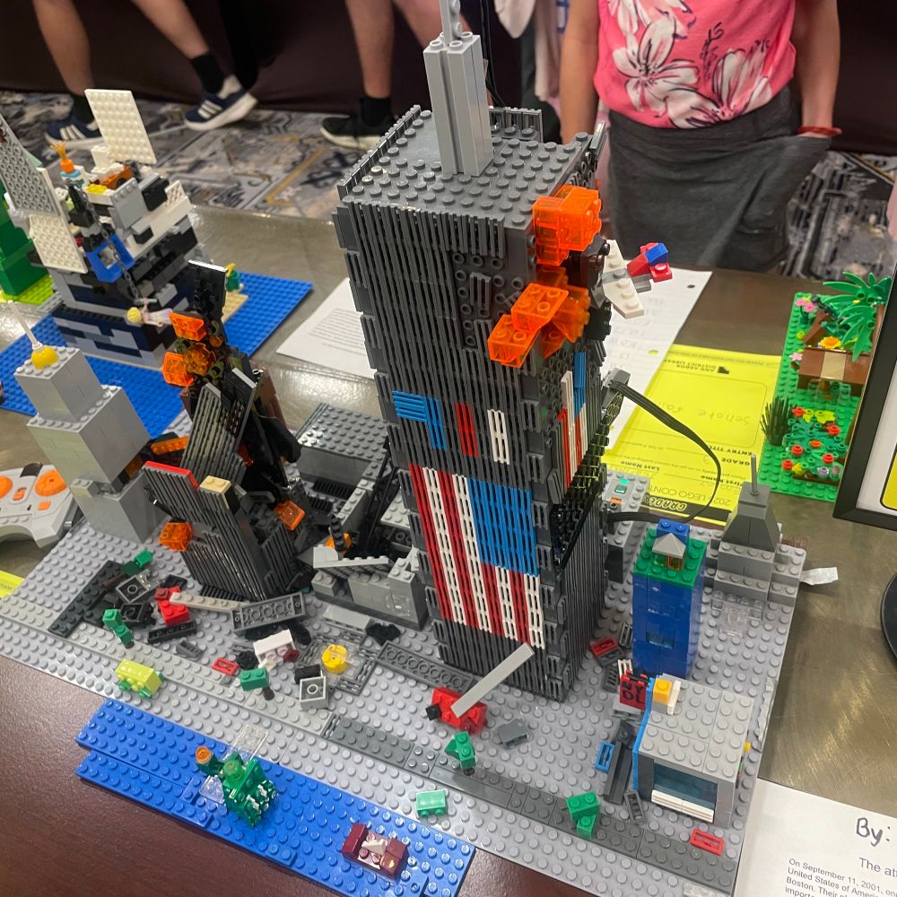 Lego 9/11. Featuring one partially collapsed tower and another one still upright. Flames protrude form the holdings and there are tiny fire trucks on the ground. The site of the standing building has American flags on it. 