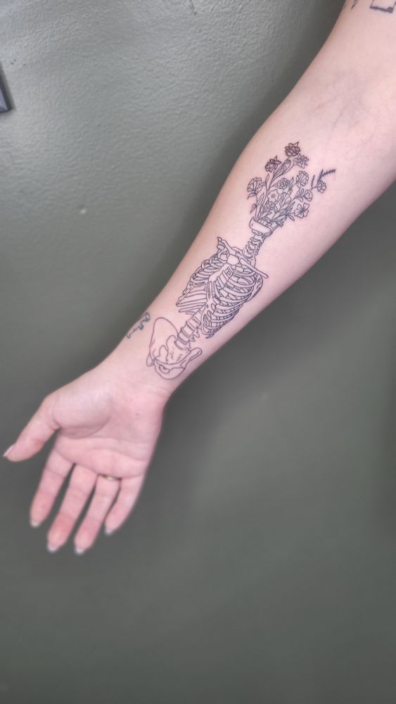A forearm tattoo of a skeletal torso with wildflowers blooming from the jawbone