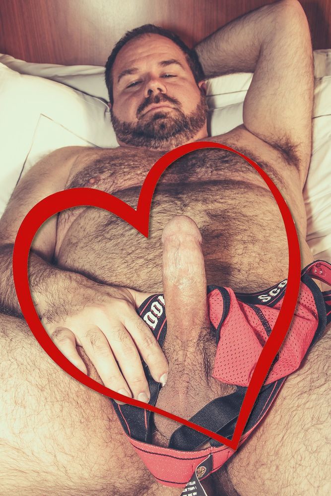Corbin Sautereau MOCubHot4U lying nude on a bed with an erection. Corbin's penis and face are visible. Corbin's left arm is cocked behind his head, exposing his hairy armpit. Corbin's right hand is gently gripping the base of his penis, and there is a red Double Scorpio jockstrap draped around it as well. There is a red heart outline edited into the photo to surround his erection.