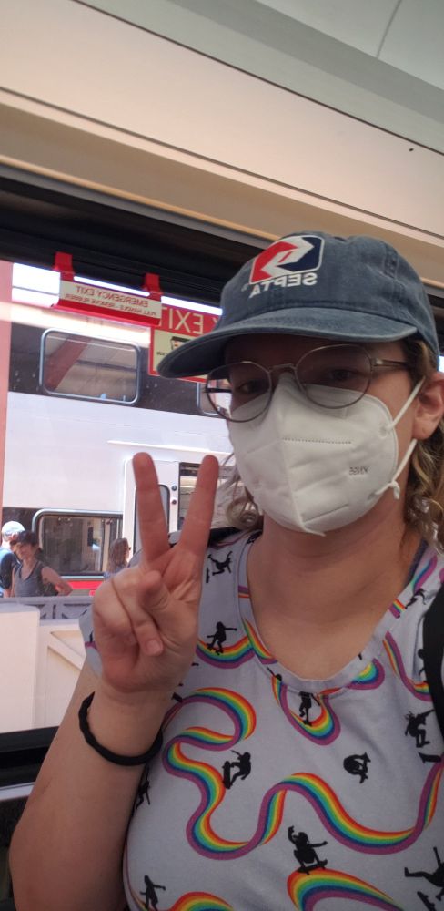 A white femme on a train, wearing a blue SEPTA hat, glasses, a white mask, and a light blue/purple shirt with girls skateboarding on a rainbow. They have a hair tie on their wrist and are holding up a peace sign. A new caltrain EMU car is visible through the window. 