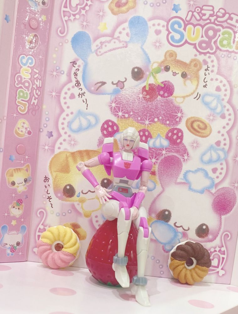 Arcee sitting on a strawberry between two donuts ,with one of her hands placed on her hip 
