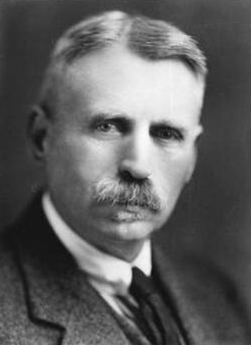 Black and white photographic, bust-length portrait of a mustachioed man with light hair and eyes. He wears a white collared short with a necktie darker than the coat and vest he also wears. He looks wistfully at a point behind the viewer's left shoulder. He like sees a pie.