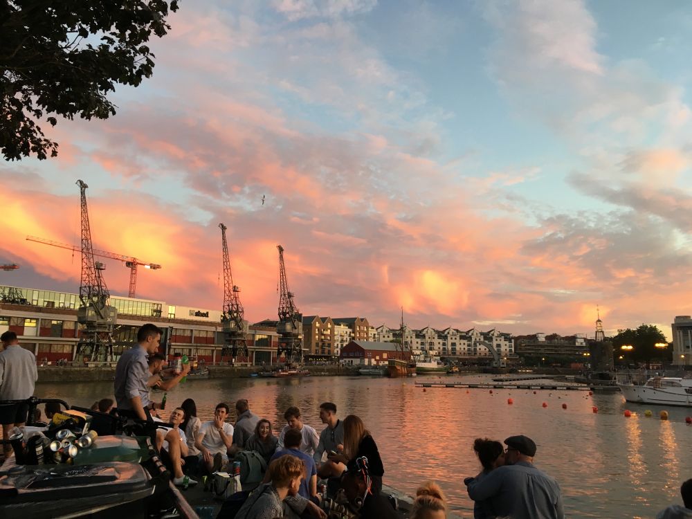 Beautiful sunset over Bristol Harbourside in the summertime. People drinking, chatting and laughing in the foreground and you can see the cranes along the waterfront in the background.