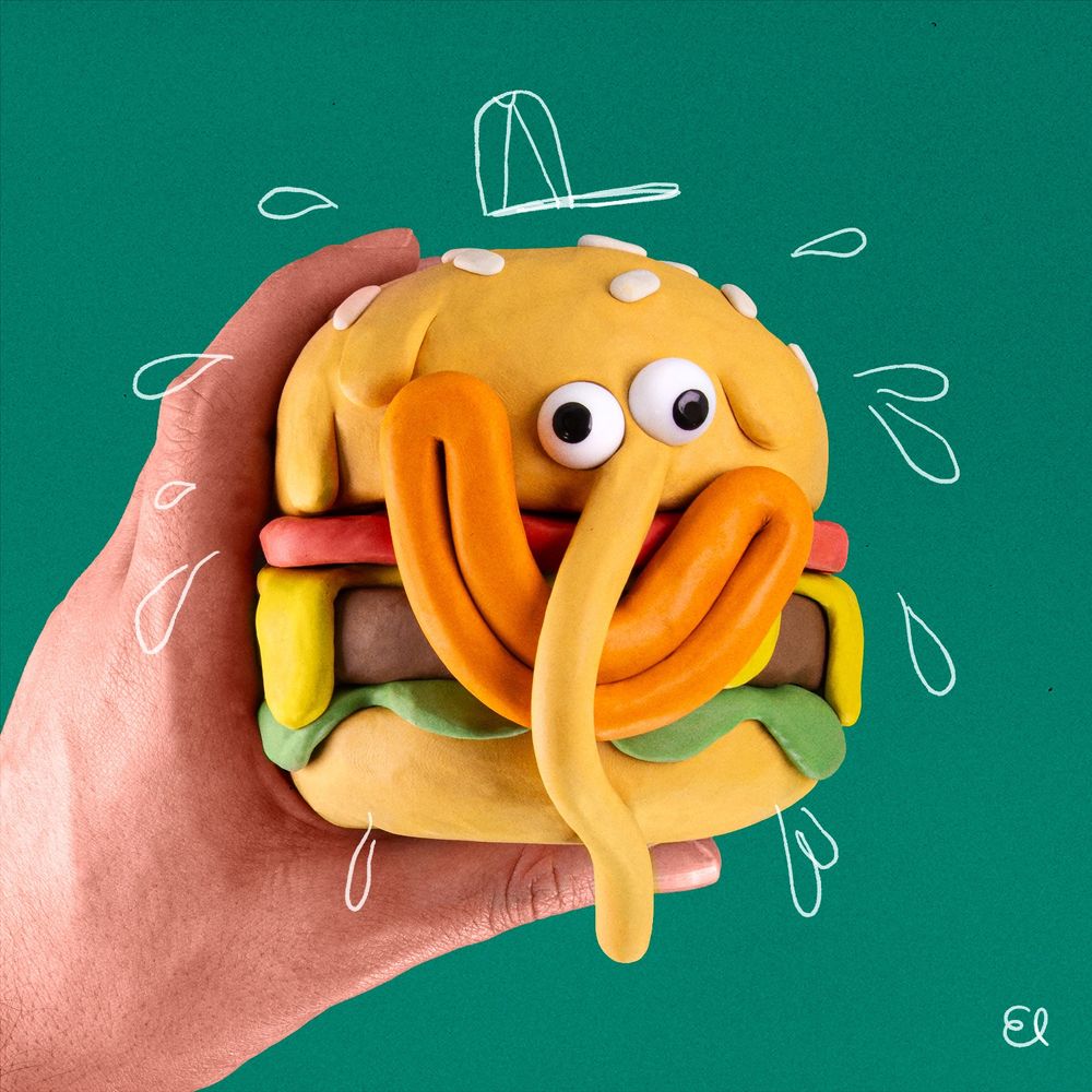 A burger sculpted from modelling clay being held up by a real hand. It has wonky eyes, big smiling orange lips and a long droopy nose.