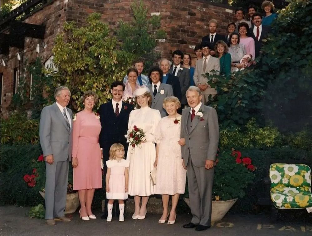 Photo of a bride and groom. And about 30 of their family and friends standing together by a set of stairs and smiling for the camera. The married couple are natalie's parents