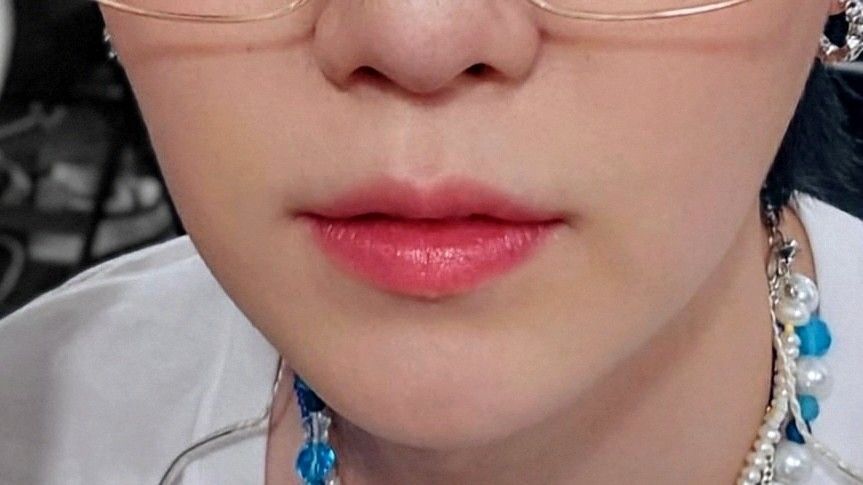 The image focus on the lower part of Yoongi's face. He's wearing a white shirt, a necklace of blue and white pearls, silver hoop earrings and glasses with large lenses. His skin is very fair, his nose so boopable and his lips pinky. The selfie was taken on the backstage of PTD on Stage in 2022. 