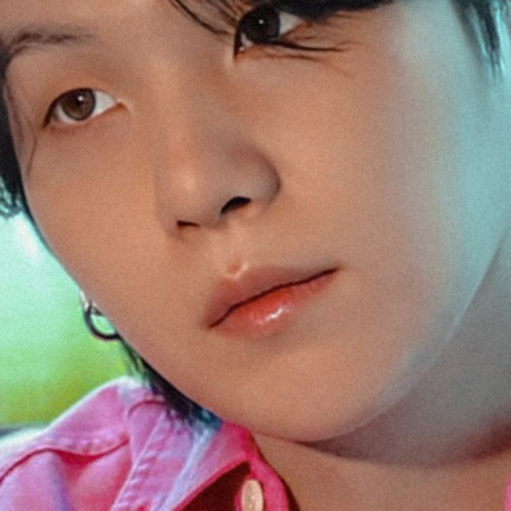 The image is a close-up of Yoongi's face highlighting his delicate features. He's wearing a pink shirt and silver hoop earrings. He's looking to his right with some hair strands gently falling on his eyes, his skin is soft and his lips pinky. The image is from the Permission to Dance Live Album.