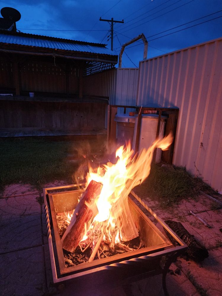 A small fire outdoors in a metal fire pit near a metal fence and a wooden bar. The sun has set, but the light has not quite faded yet.