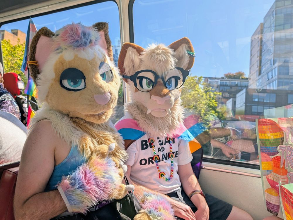 two Siamese cat fursuiters on the monorail, clear sky and a few buildings in the background 