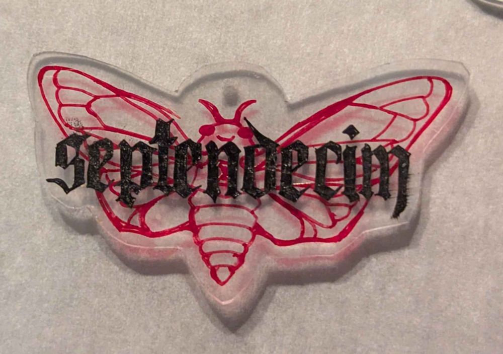 a close-up photo of a clear plastic shrinky dink charm, showing a cicada in red line art. the Latin word "septemdecim", seventeen, is written in black ink in a Gothic style.