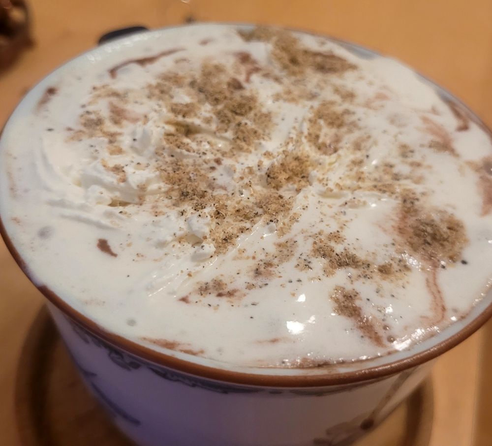 Cup almost overflowing with cocoa, topped with cream and a sprinkling of cardamom