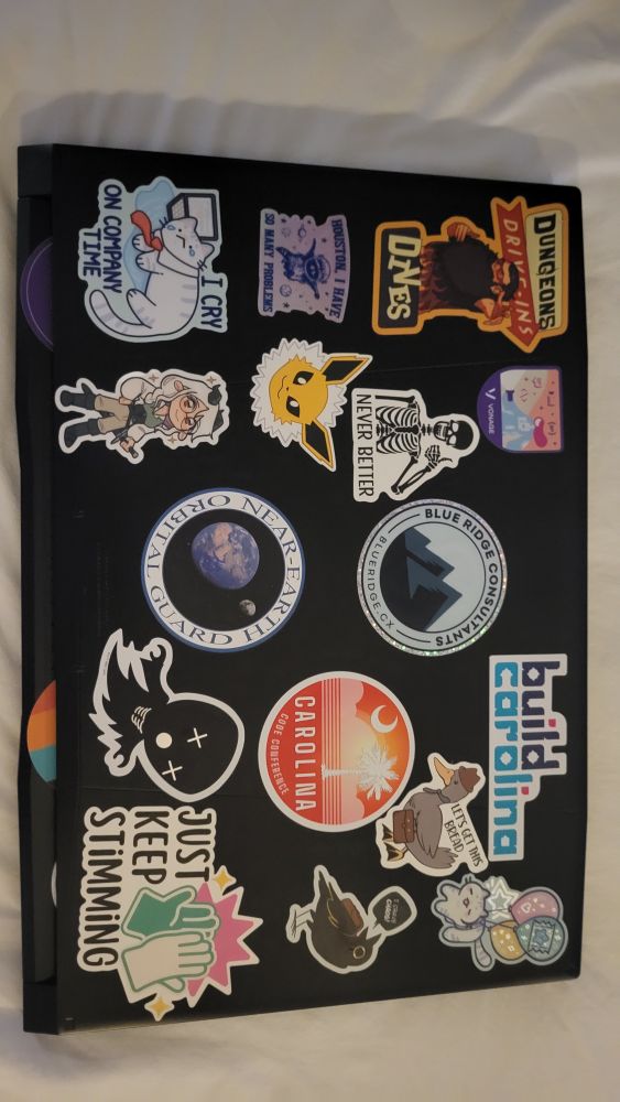 A laptop with stickers. A Dungeons Drive-ins and dives sticker with Sensei.  A raccoon in space that reads "Houston I have so many problems", A cat crying on a laptop that reads "I cry on company time", A Vonage dev sticker, a skeleton with a thumbs up reading "never better", a Jolteon head, a character from the webtoon Realta, a blue ridge consultants sticker, a Near Earth Orbital Gaurs sticker from K.B. Wagers NEO-G series, a Build Carolina sticker, a duck in a hat that reads "let's get this bread", a Carolina Code Conference sticker with a South Carolina flag motif. A wired ghostie thing a cat with balloons. Walden, a crow from Realta, wearing a backpack and saying "I crave chaos" and a Autism support sticker that reads "just keep stimming"