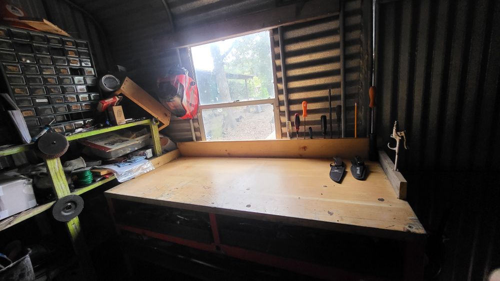 A clean workbench with 2 knives and screwdrivers at the ready. I window overlooks a tree covered yard. Varies items sit disorganized on a shelving unit. 
