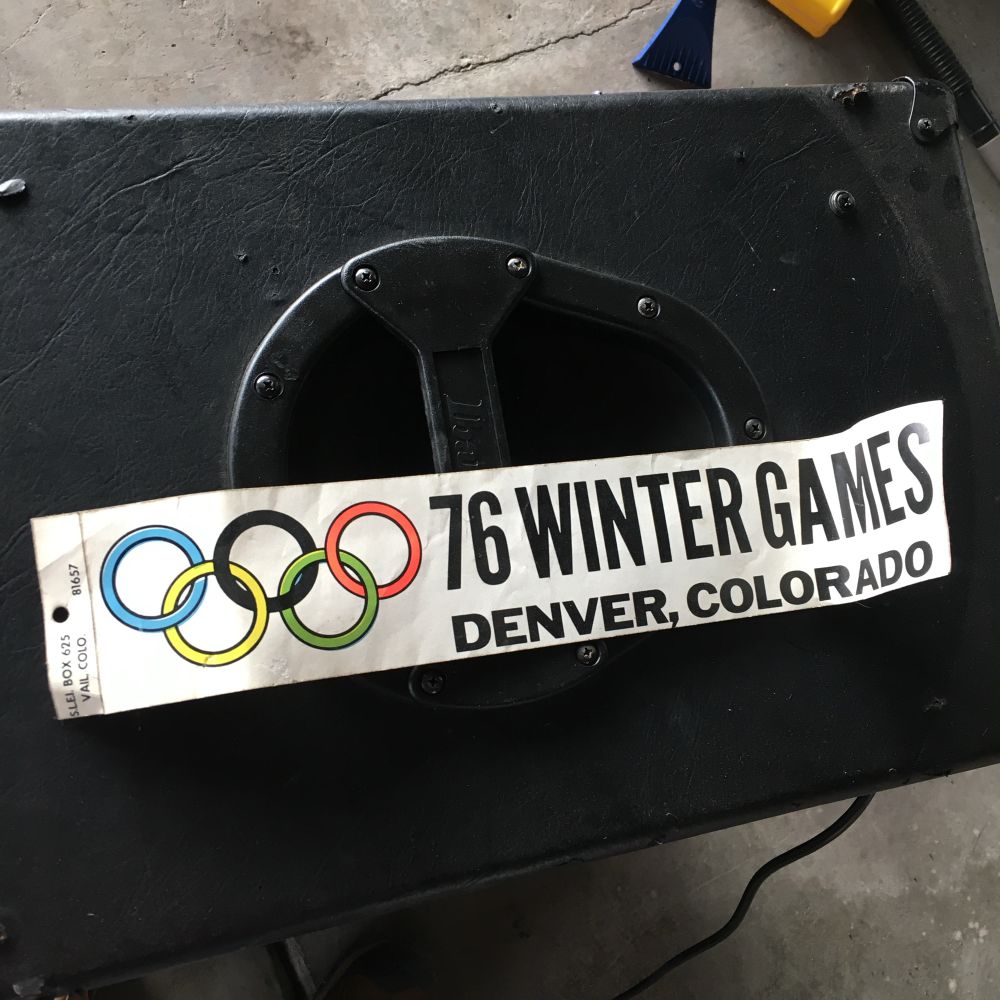 An old bumper sticker atop a large guitar amplifier in a grungy garage. The sticker displays the Olympic rings and says “76 Winter Games / Denver, Colorado